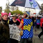 Wonder Woman protesting with umbrella that is painted with a flower and "They tried to bury us they didn't know we are seeds"