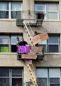 Dudes on fire escape with signs: FACTS RULE, and "Make Signs Great Again"