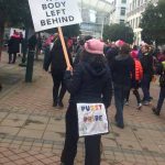 back of protestor with booty flap reading "Pussy Power" and a sign for disability rights: No Body Left Behind