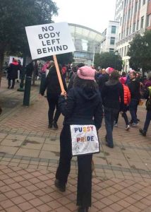 back of protestor with booty flap reading "Pussy Power" and a sign for disability rights: No Body Left Behind
