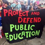 woman dressed as Wonder Woman holding huge banner: Protect and Devent Public Education