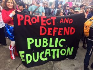 woman dressed as Wonder Woman holding huge banner: Protect and Devent Public Education