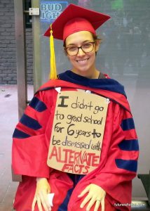 Woman in cap and gown with sign: I didn't go to school for six years to be dismissed with alternative facts