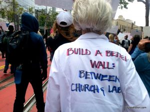 Woman's lab coat reads: BUILD A WALL BETWEEN CHURCH AND LAB.