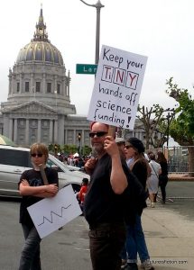 A protest sign with "Keep your TiNY hands off science funding!" where TiNY are elements from the periodic table.
